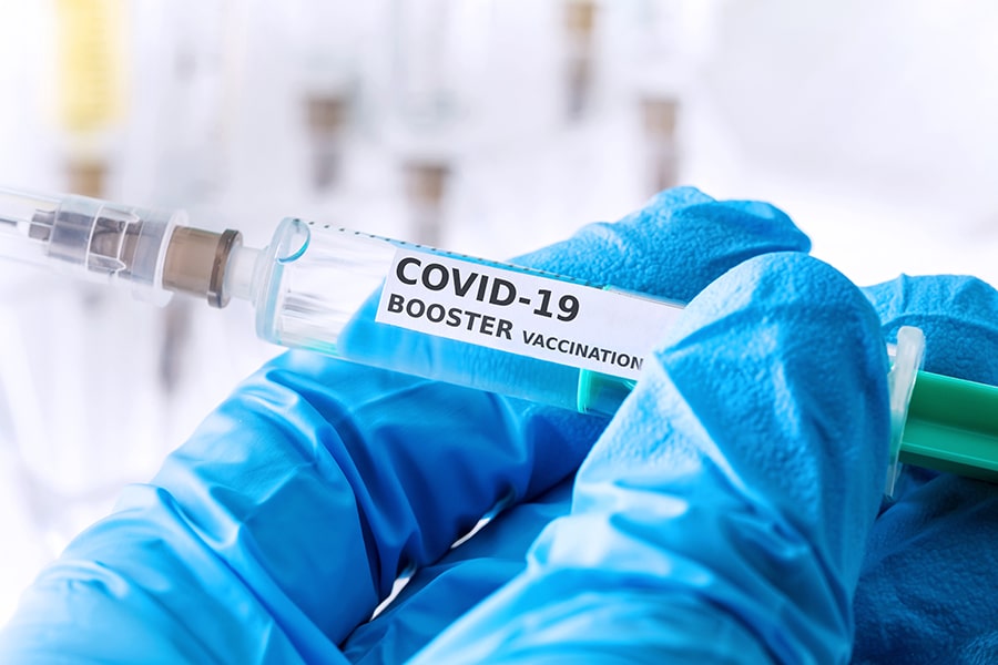 a gloved hand holding a syringe labeled with COVID-19 BOOSTER VACCINATION