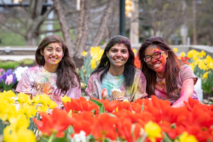 students covered with colorful chalk after Holi pose near flowers