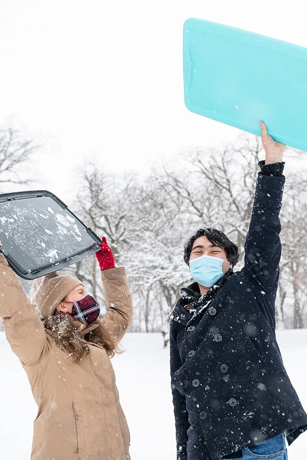 Two masked graduate students stand on a snowy hillside, laughing and holding lunch trays, used as makeshift sleds, in the air.