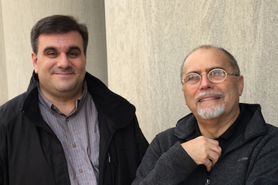 An outside shot of two smiling men in front of a tan, nondescript building. Both are dressed in light black jackets