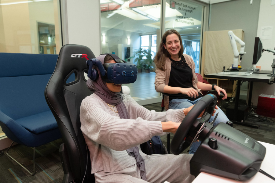 woman wearing a VR headset driving a car simulator with a profsesor smiling
