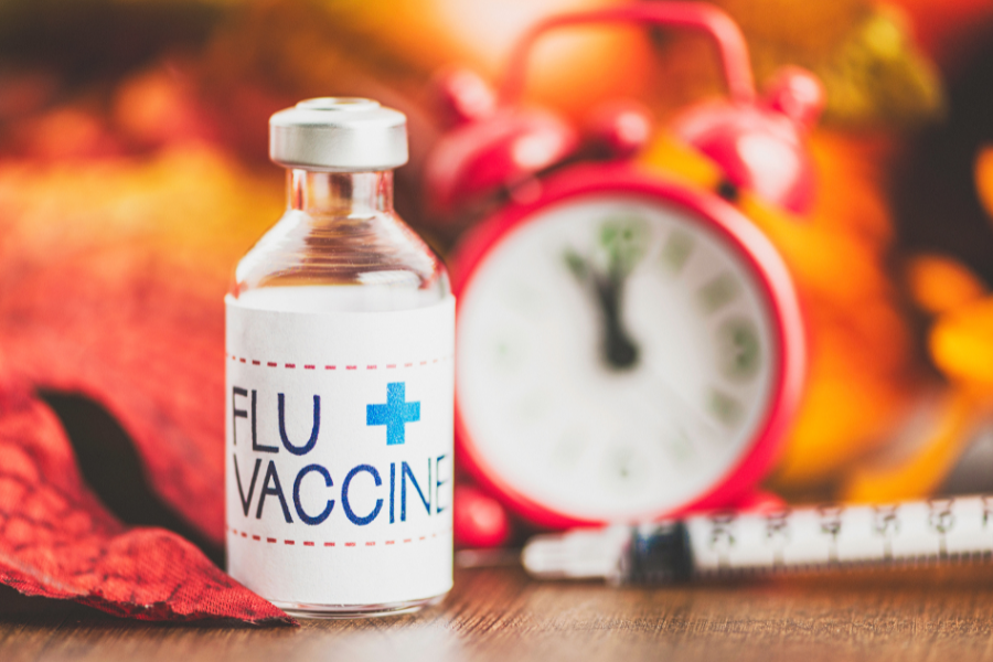 image of a flu vaccine vial next to a clock and syringe, surrounded by leaves turning shades of orange, yellow, and brown. 