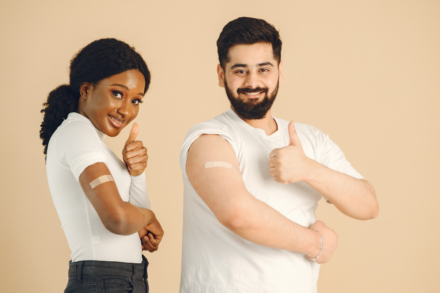 An image of two people from torso to head, they are pulling up their sleeves to show bandaids on their arms 