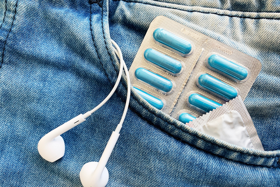 a close up image of a jeans pocket, there is a condom package, pills, and headphones sticking out of the pocket