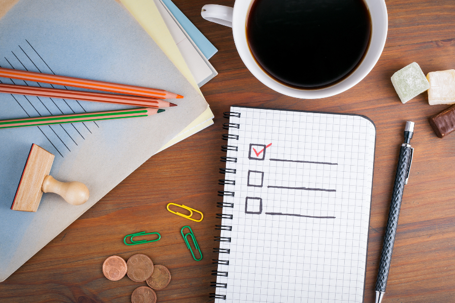 A notebook with a check list sits upon a table with a pen, a coffee mug, colored pencils, paper clips, and papers.