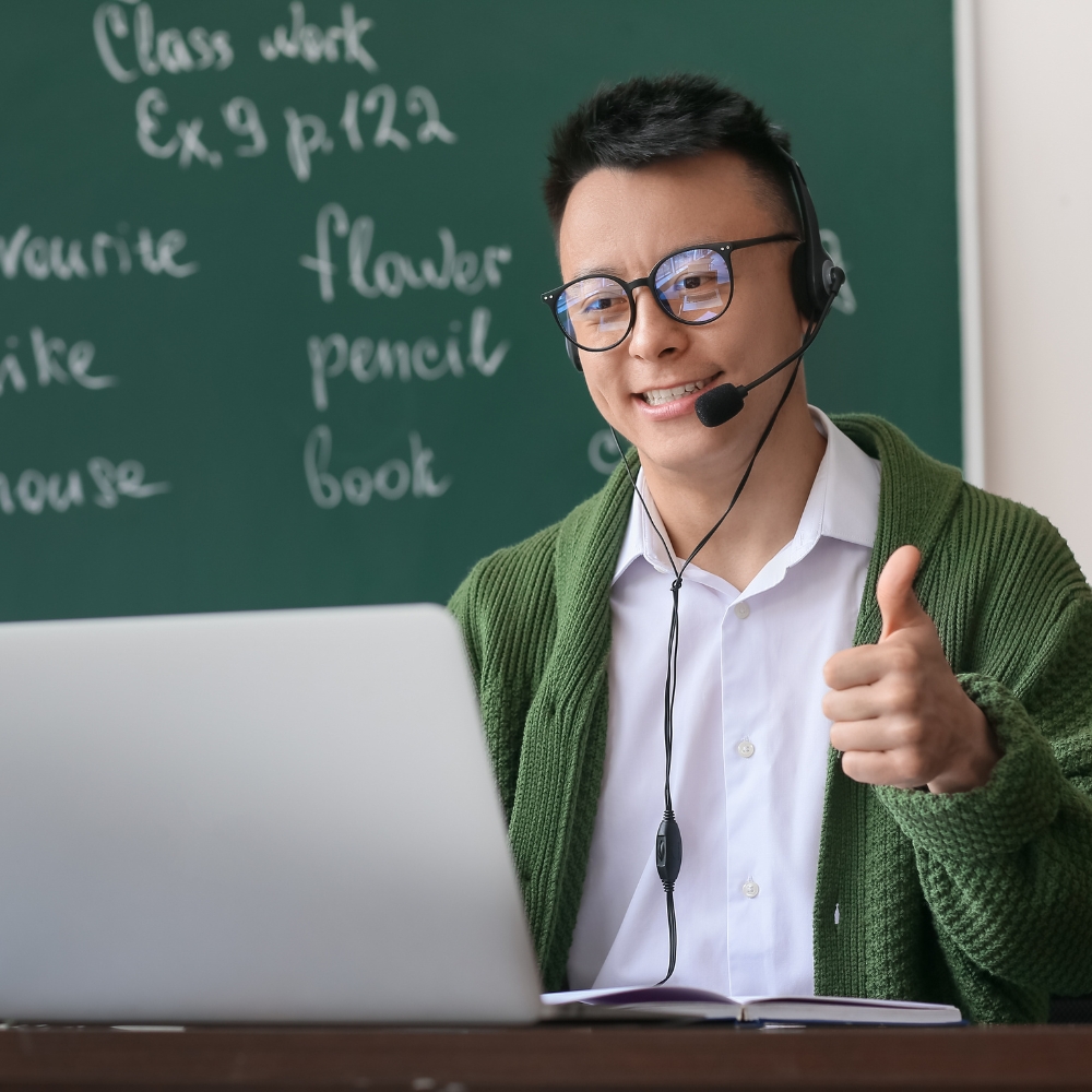 Teacher with green chalkboard background gives thumbs up sign. He is sitting at a teacher's desk in front of a laptop with his computer screen open.