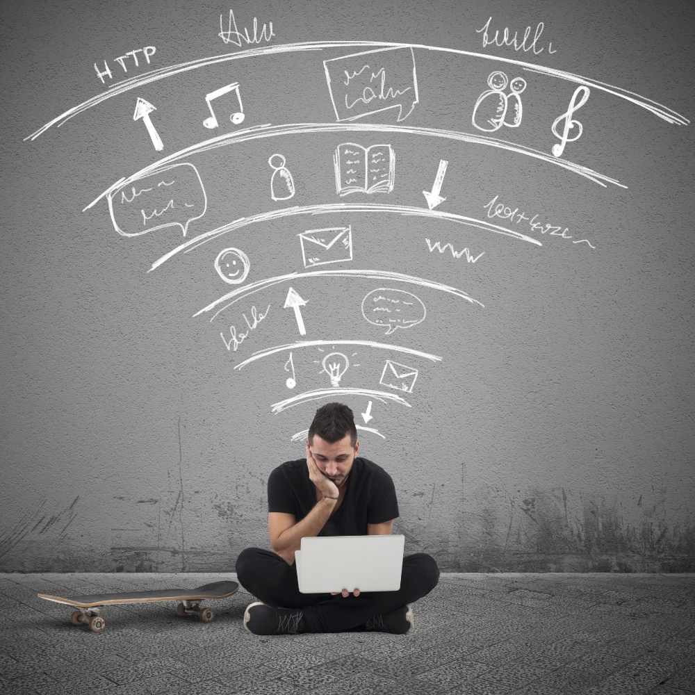 A student sits cross-legged with a laptop on his lap; gray background; words and images suggesting wifi connection are on the wall behind him.