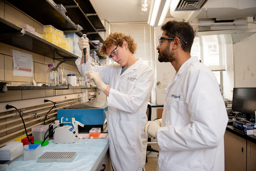Two scientists in white lab coats and safety goggles perform experiments in a chemistry lab.
