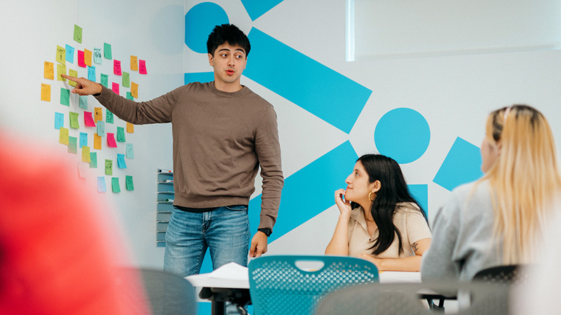 A student presents ideas from a wall covered in sticky notes arranged in a heart shape
