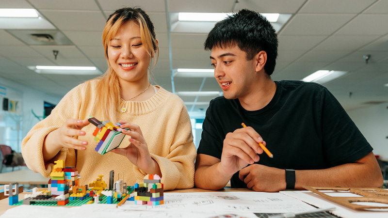 Two students build a colorful structure with LEGO bricks while reviewing sketches on the table