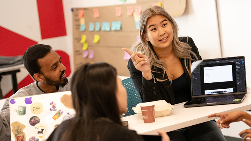 A student presents her laptop screen to classmates seated around a table lined with sticky notes