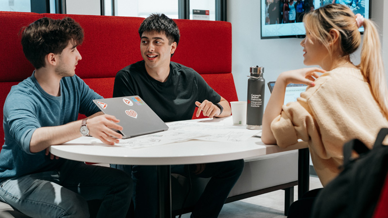 Three students talk together on a lounge seat, one holding a laptop covered in stickers