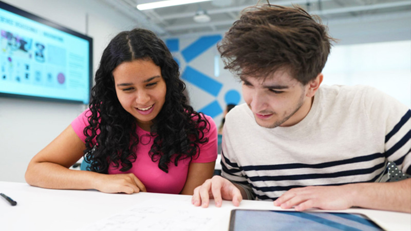 Two students review notes together at a table with a data visualization on a screen nearby