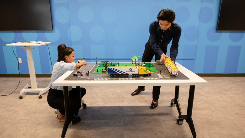 Two students build a model city with LEGO bricks and trucks on a large worktable