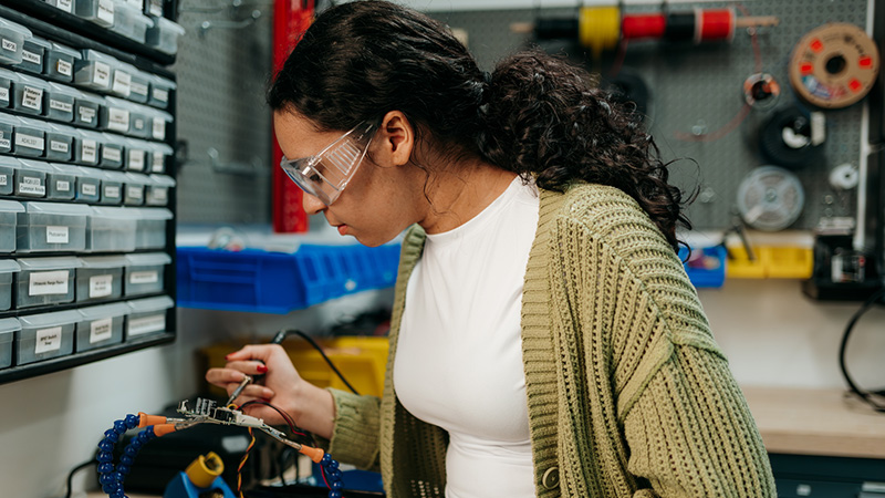 A student wearing safety glasses solders an electronic component at a workbench