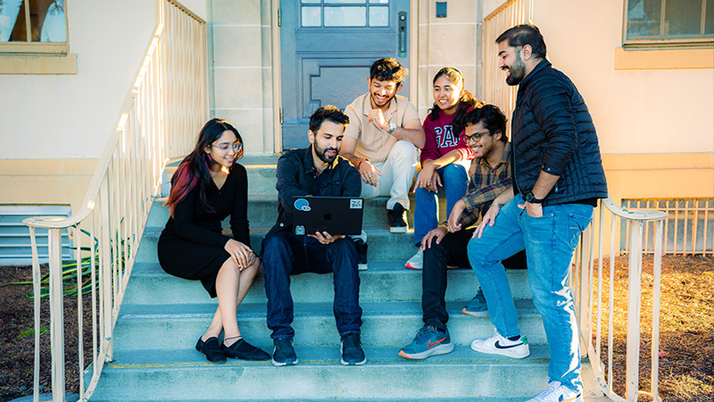 Three students stand beside a printed pitch poster for a startup called PulseBoard