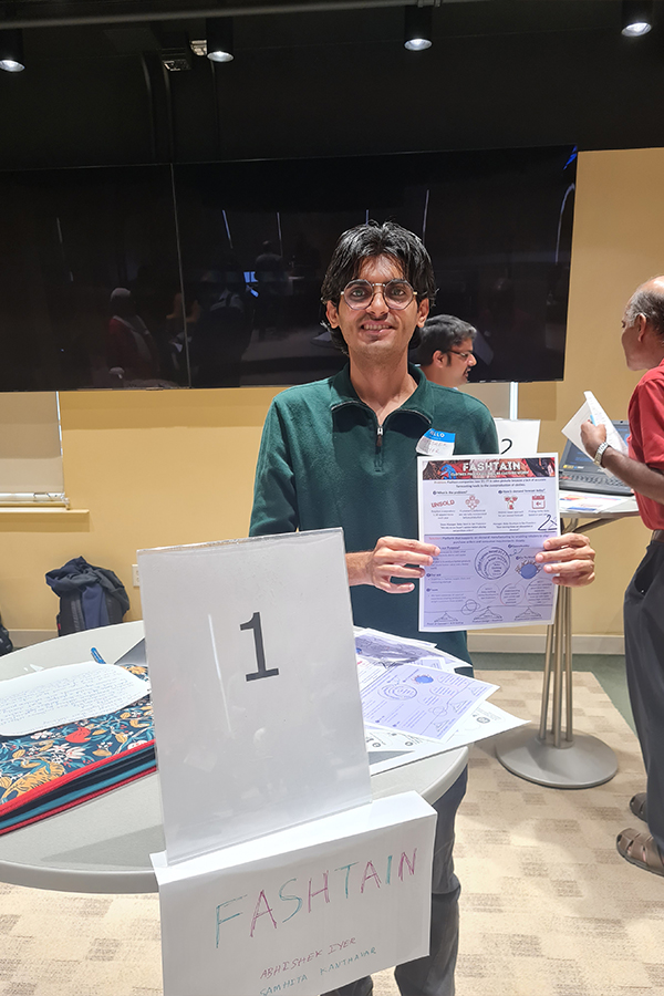 Abhishek is standing at a circular table holding a flyer with information about his startup, Fashtain