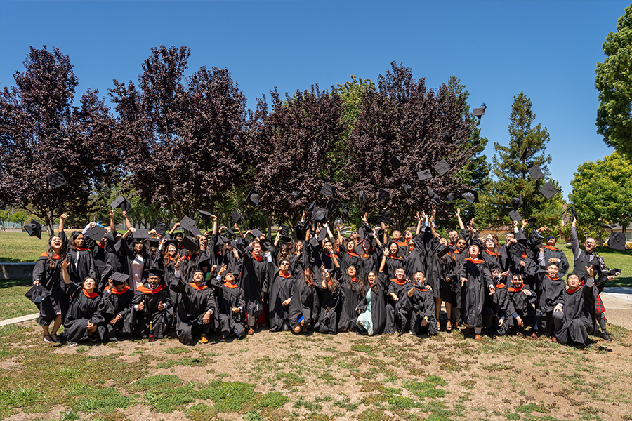A group of students and alumni all pose together for a photo. They are wearing black graduation gowns with orange hoods. The group is all throwing their graduation caps in the air in celebration