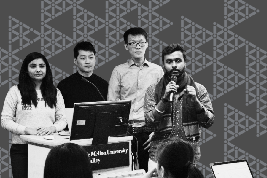 A group of students stand at the front of a classroom with a large screen showing a presentation behind them. One of the students stands with a microphone in hand and is speaking and gesturing while presenting.