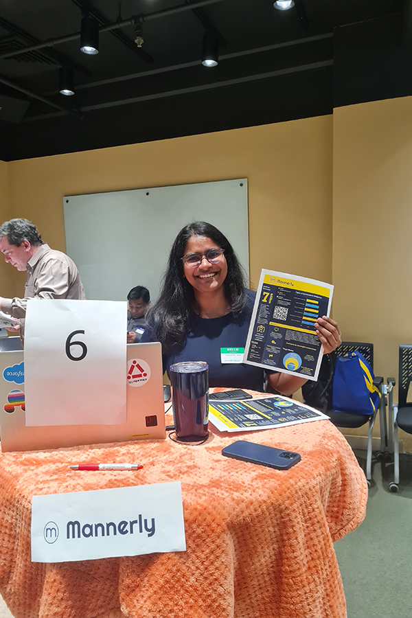 Pranathi stands at a circular table holding pamphlets about her startup, Mannerly