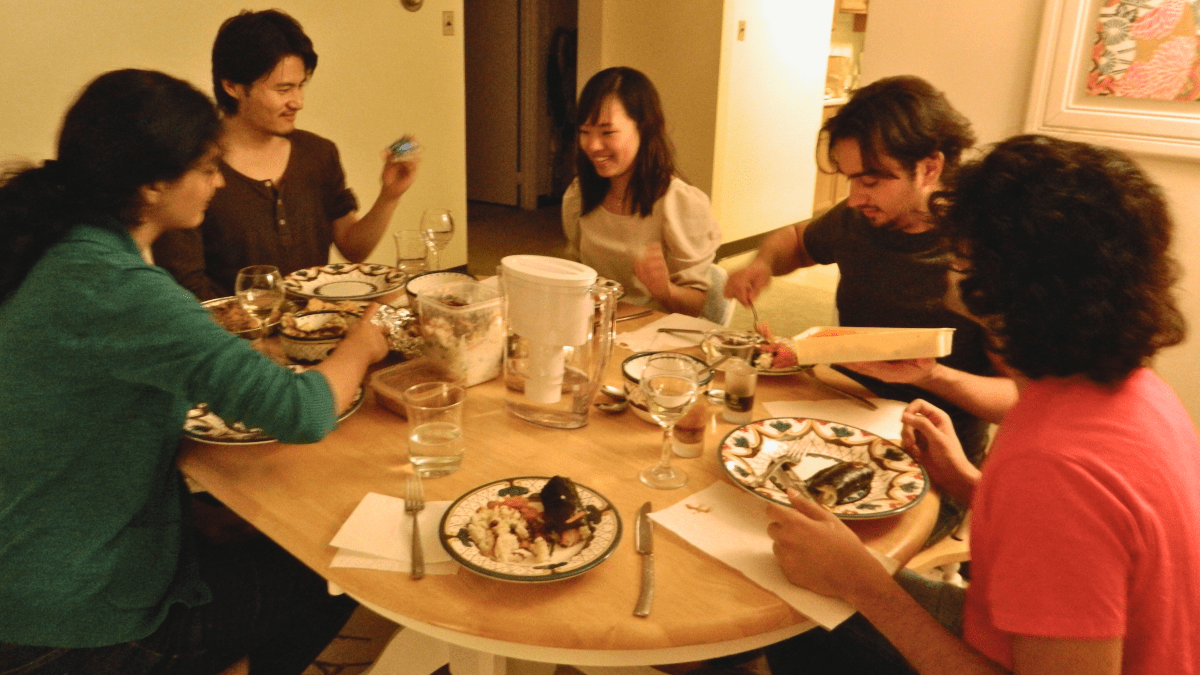 Seated at an oval-shaped table, five people enjoy dinner as they talk.