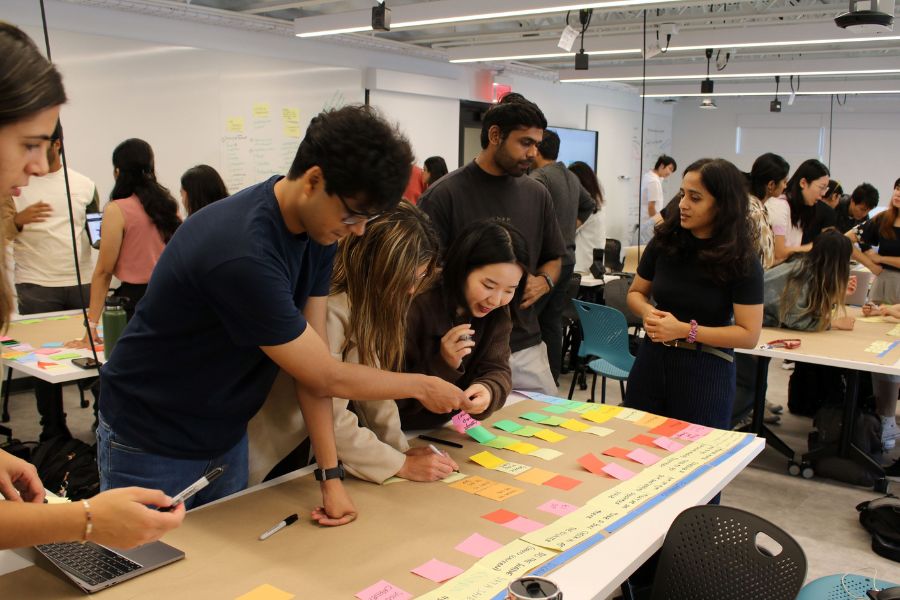 Several groups of students are spread out across a classroom, each working at a table. The main group has a grid of colorful sticky notes organized by color.