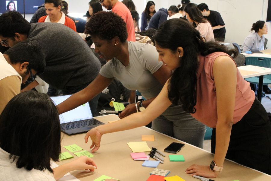 Two students reach across their project desk to move green sticky notes to another section of their map. Multiple students, working in their groups, are visible in the background.  