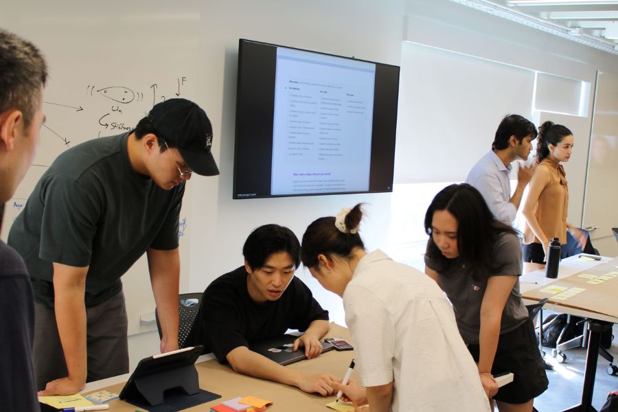 To the left of a flatscreen TV depicting instructions, a group of 5 students huddles over a desk as one begins writing; two students from another team are in the background.