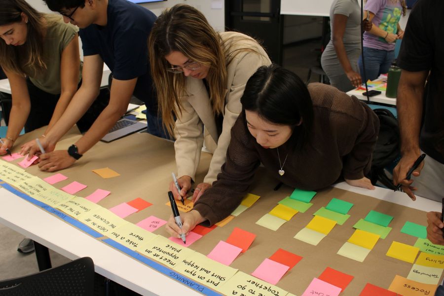 A rainbow of blank sticky notes fills the right side of the table, organized in 6 rows by color – pink, red, light yellow, yellow, green, and teal – across more than 10 columns. Four students, on the team of 6, are visible and have begun populating the top row. 