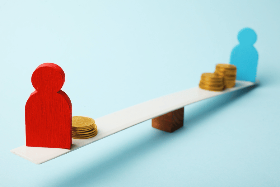 Against a pale blue backdrop, a wooden block figure in red with 6 gold coins sits on the raised end of a seesaw, facing a sky blue wooden block figure with 3 times the gold coins. 