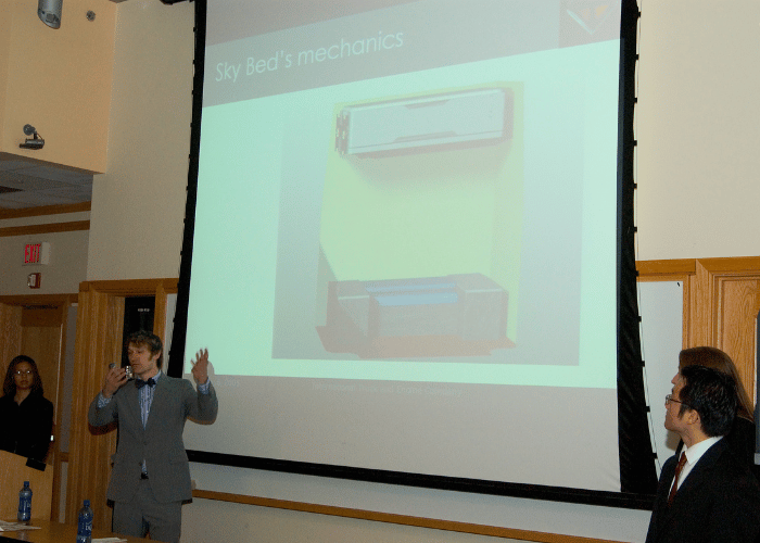 Pontano, in a grey suit and bow-tie, presents in front of a large pull-down projection screen depicting the solution.