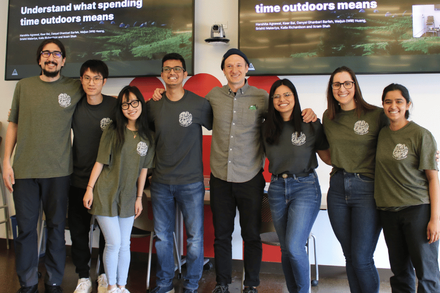 The seven students of Team Ignik and Peter Pontano pose in the front of the classroom with their Phase 2 presentation slide - titled "Understand what spending time outdoors means" - on two flat screen TVs above the group.