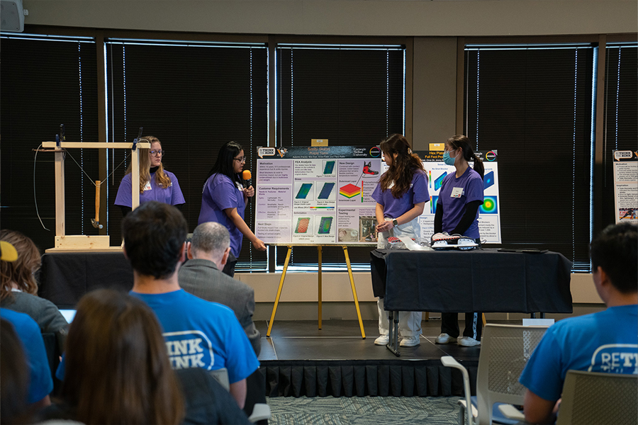 a team of students wearing purple shirts stands on a stage. the students are in front of a large poster board containing their design for the project. one student points to the poster board and the others are looking along at them.