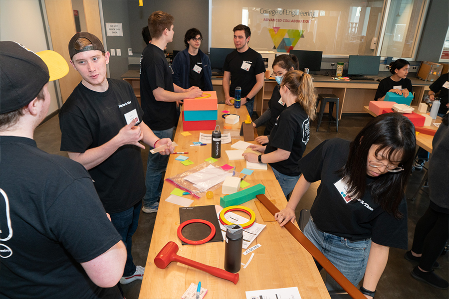 four students wearing black shirts are at a tall wooden table. on the table, various materials like plastics, foam, and paper are spread out. students are conversing with one another and inspecting the materials.