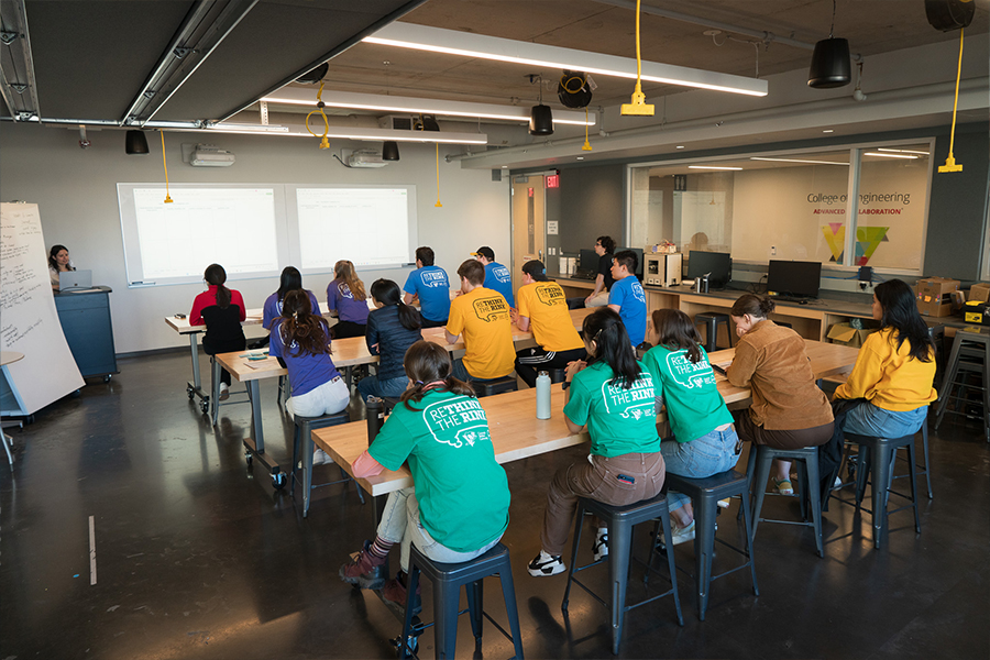 students wearing different colored shirts sit at three long tables and listen to a speaker at the front of the room