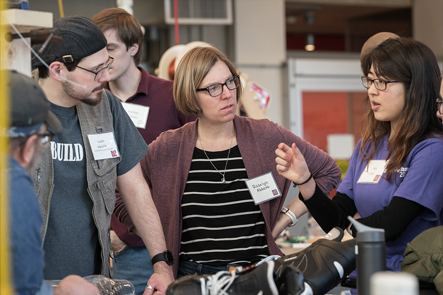a student wearing a purple shirt explains her team's design to two mentors. the design, a hockey skate with a special shot blocker, is laying on the table in the forefront of the image.