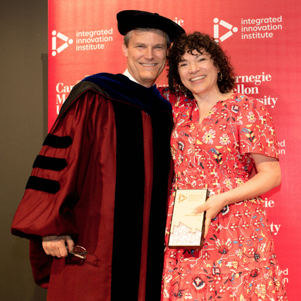 iii Director Peter Boatwright, dressed in doctoral regalia, smiles alongside Minette Vaccariello as they pose in front of the Institute's step and repeat branded backdrop with her alumni changemaker award