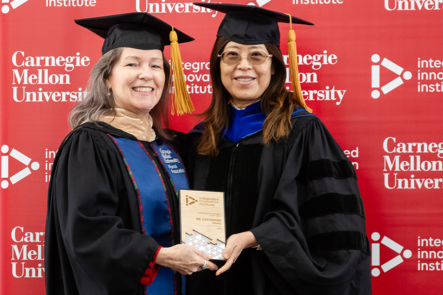 Two women stand against a red backdrop in graduation regalia, they hold an award between them and are smiling at the camera