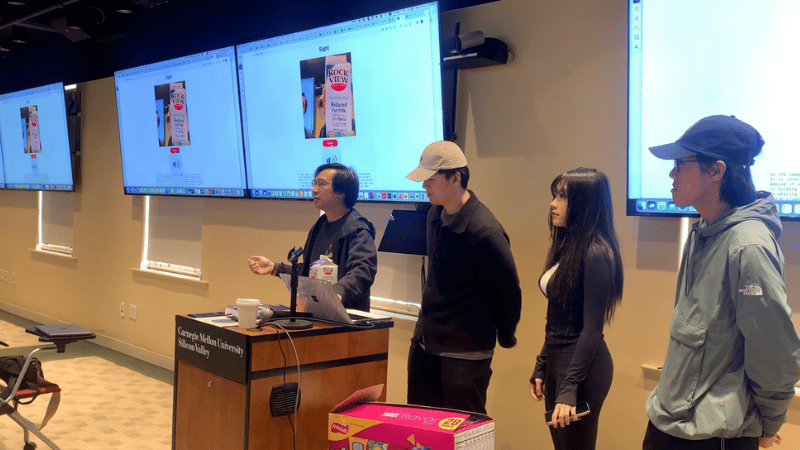 Four students stand side-by-side to the right of the podium as one student demonstrates their product via a video in the presentation. 5 large monitors display the students' presentation