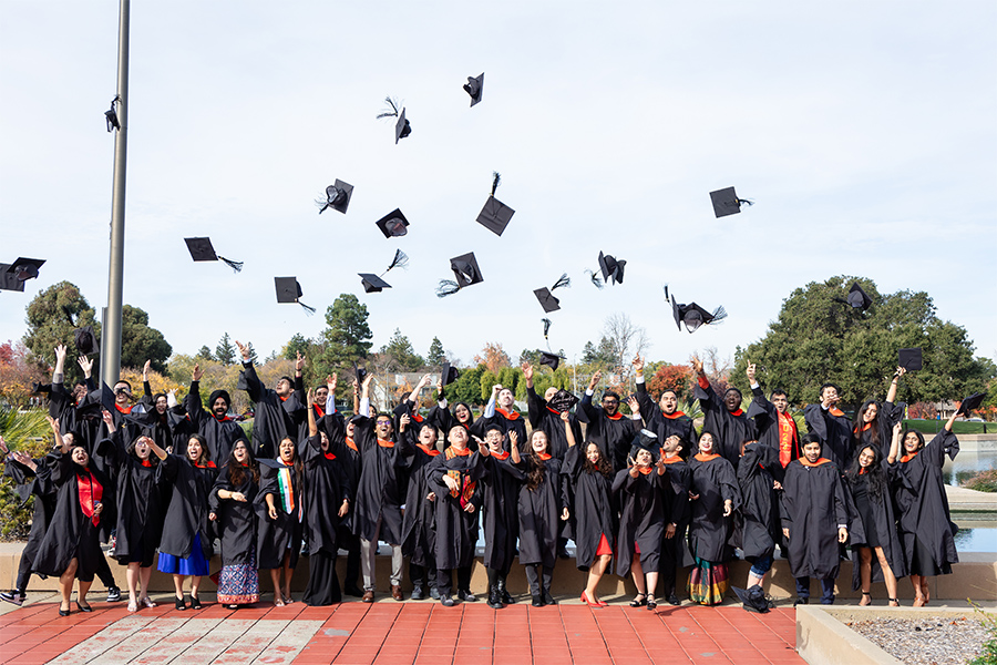 A group of graduates stand in a group wearing their graduation gowns and regalia. They are cheering excitedly as they toss their caps into the air