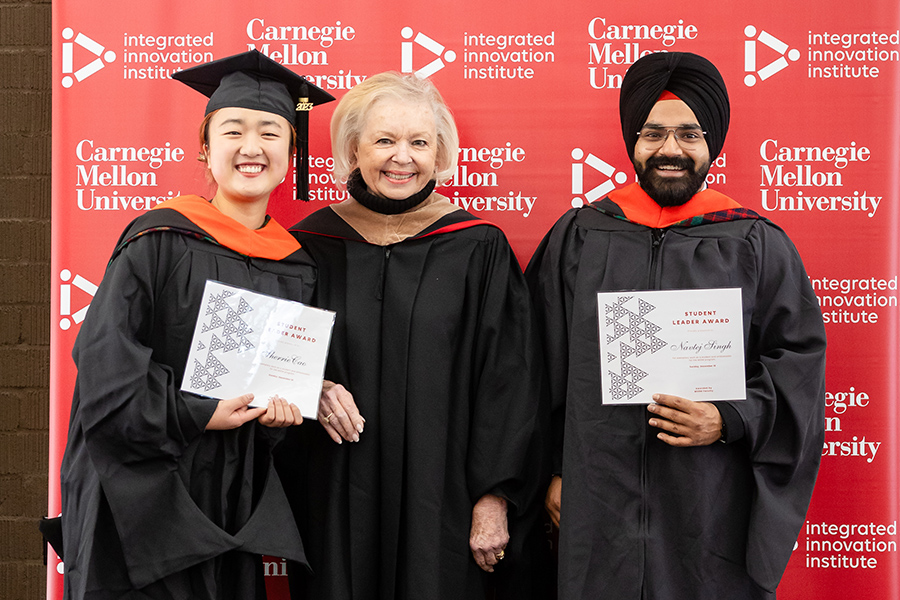 Two students stand beside their professor against a red backdrop, all dressed in graduation regalia. The students each hold a certificate.