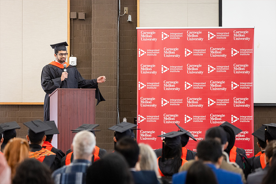 A student dressed in graduation regalia addresses fellow graduates from a podium on a stage