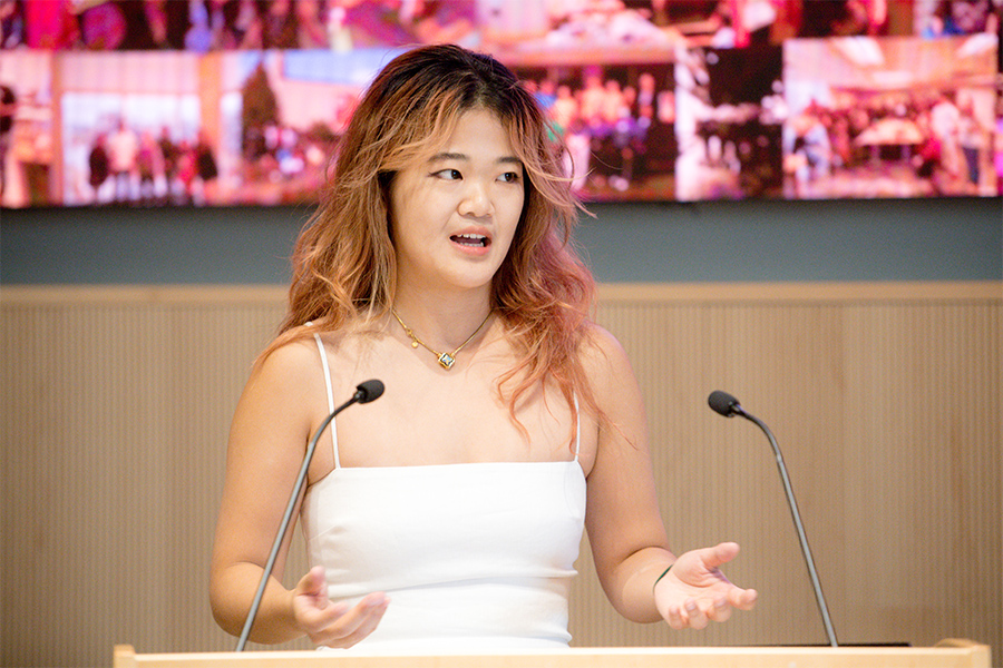 A woman in a white dress stands at a podium. She is mid-speech and holding her hands up while talking.