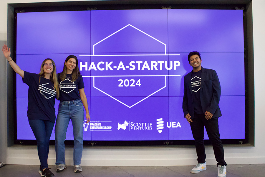 Three MIIPS students pose for a photo in front of a digital screen that reads "Hack-a-Startup 2024"