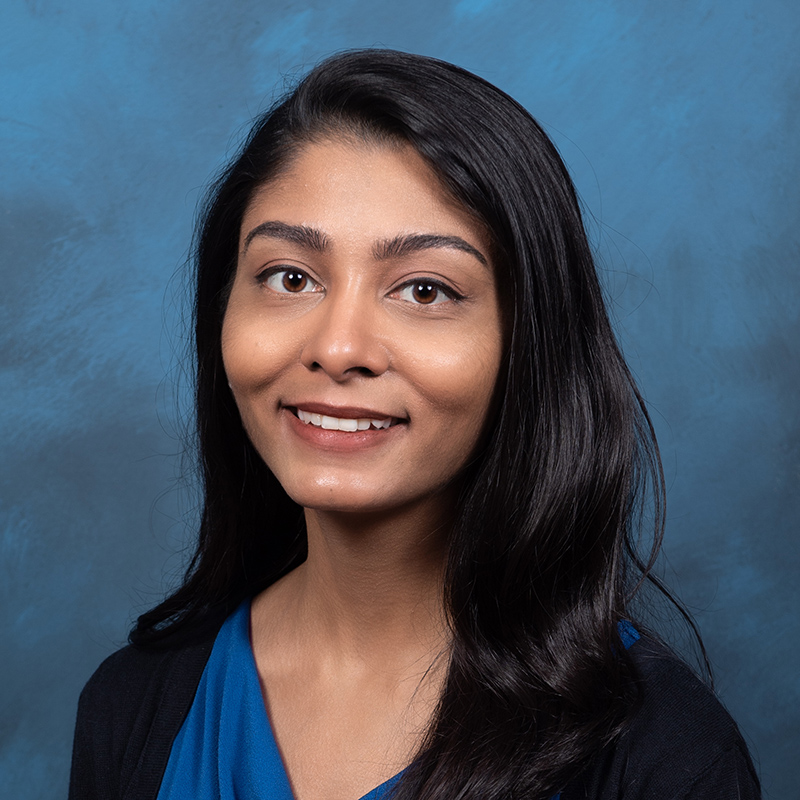 A woman from India with long brown hair in a blue v-neck shirt against a slate-blue background