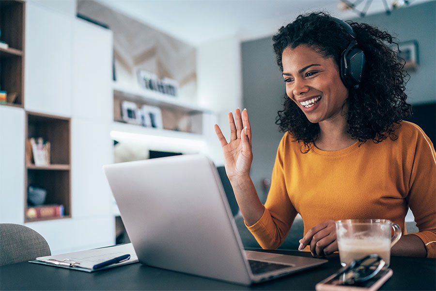 A woman is sitting at her desk and is participating in a video call. She has her hand raised and is smiling.