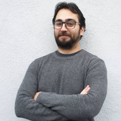 Tomas Alvarez who has dark hair, glasses, and a beard poses with arms crossed against a white wall