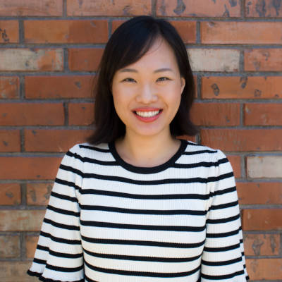 Hannah Kim who has chin-length brown hair smiles wide as she poses against a brick background