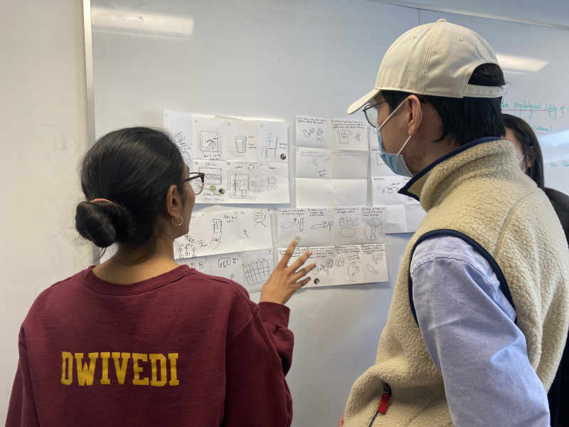 Dwivedi and two classmates stand in front of a large white board, discussing product ideas, which are depicted across five pieces of white paper.