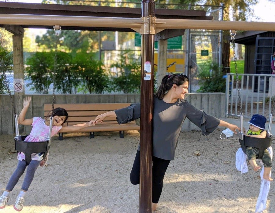 Divya stands at the center, reaching out to hold each child's hand. The children are each in a swing at the park.